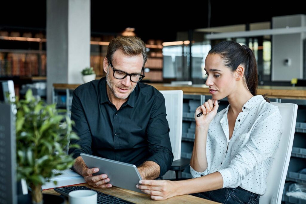 A woman and a man working together in an office, looking a at a tablet and thinking